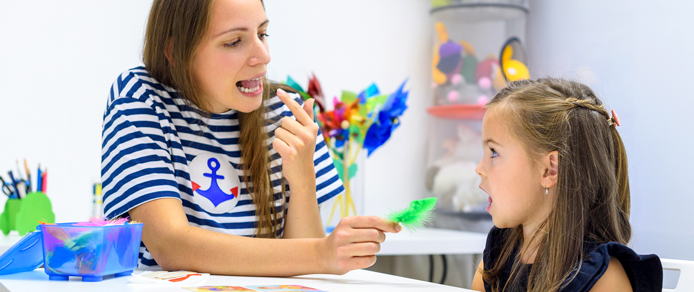 A teacher in a blue and white striped shirt sits next to a young girl at a classroom table. The teacher points to her own mouth while making letters sounds while the girl mimics her.