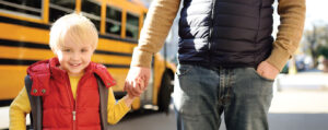 Young boy holds his father's hand near yellow school bus on background. Kindergarten transition.