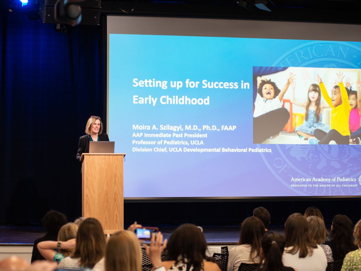 Dr. Moira Szilaygi speaking in front of a crowd with a projected screen to her right titled: Setting up for Success in Early Childhood.