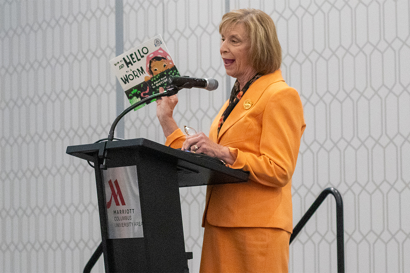 Ohio First Lady Fran DeWine holds the children's book "How to Say Hello to a Worm" in her right hand as she speaks into a microphone behind a podium at the 2023 Crane Symposium on Children.