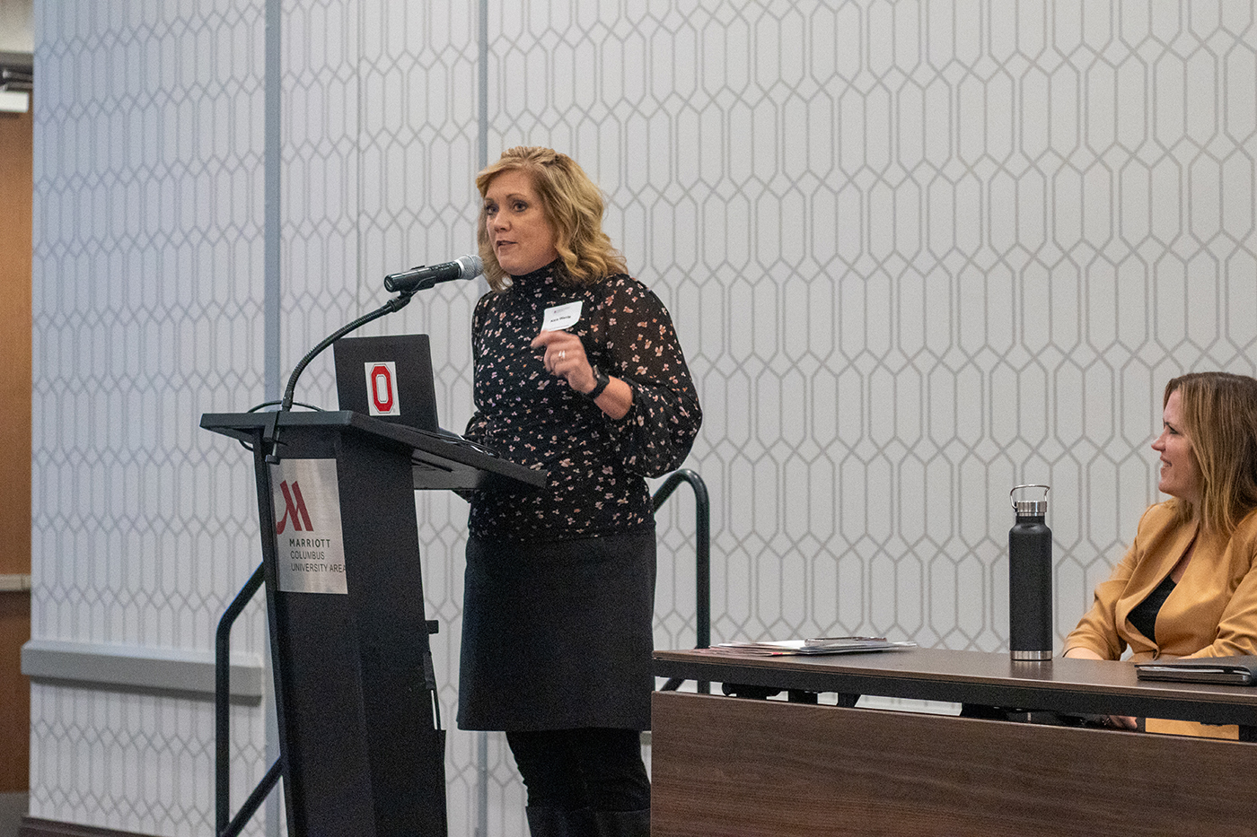 Kara Wente, director of the Ohio Department of Children and Youth, stands behind a podium and speaks into a microphone attached to the podium during the 2023 Crane Symposium on Children. To the right, Dr. Laura Justice, Crane Center executive director, sits behind a table and listens.