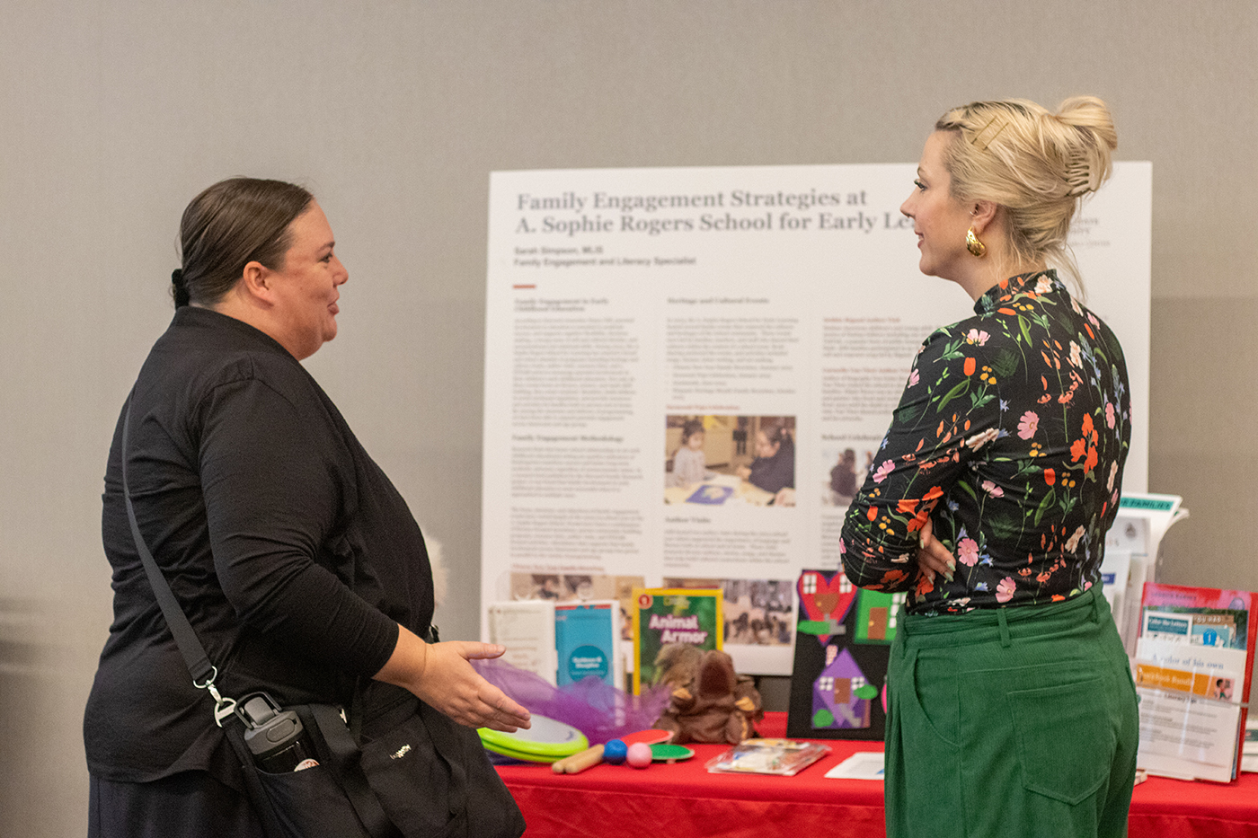 Two people speak in front of a poster display at the 2023 Crane Symposium on Children. The poster display describes family engagement strategies at Ohio State's A. Sophie Rogers School for Early Learning, and in front of the poster is a table on which are children's books, toys and informational handouts.