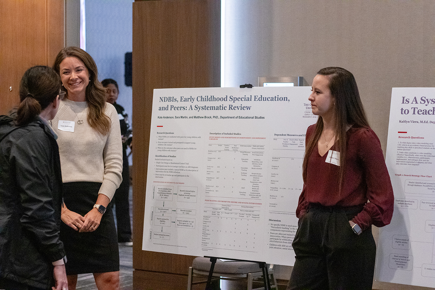 Crane Center graduate research associates Kate Anderson, left, and Kaitlyn Viera, stand next to posters discussing their research during the 2023 Crane Symposium on Children. Anderson is smiling while listening to a symposium attendee immediately in front of her, while Viera looks on.