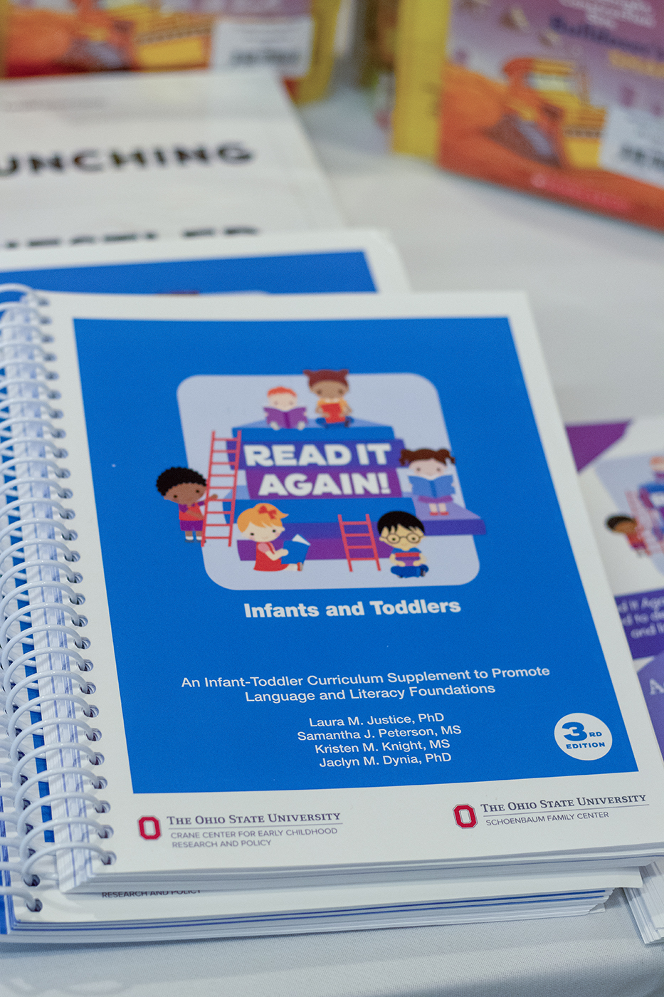 A stack of booklets titled "Read It Again! An Infant-Toddler Curriculum Supplement to Promote Language and Literacy Foundations" rest on a table. The booklets are spiral-bound and have a blue cover with a cartoon illustration showing a variety of small children reading books.