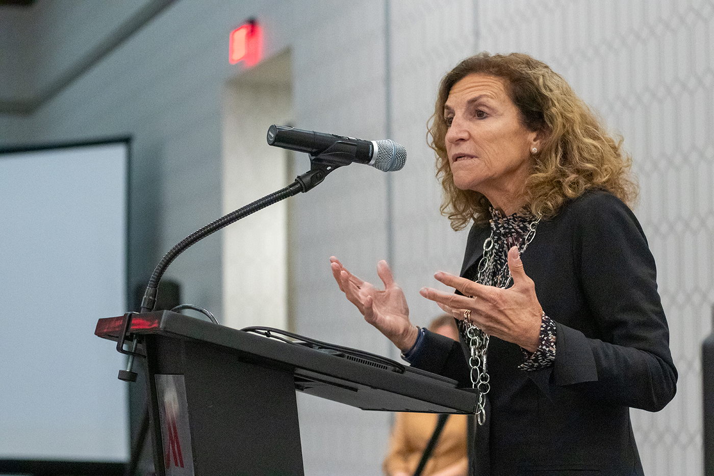 Tanny Crane, president and CEO of Crane Group, speaks at the 2023 Symposium on Chlldren. Crane stands behind a podium, speaking into a microphone attached to the podium. Crane holds her hands outward, with arms bent at the elbows, as she speaks.
