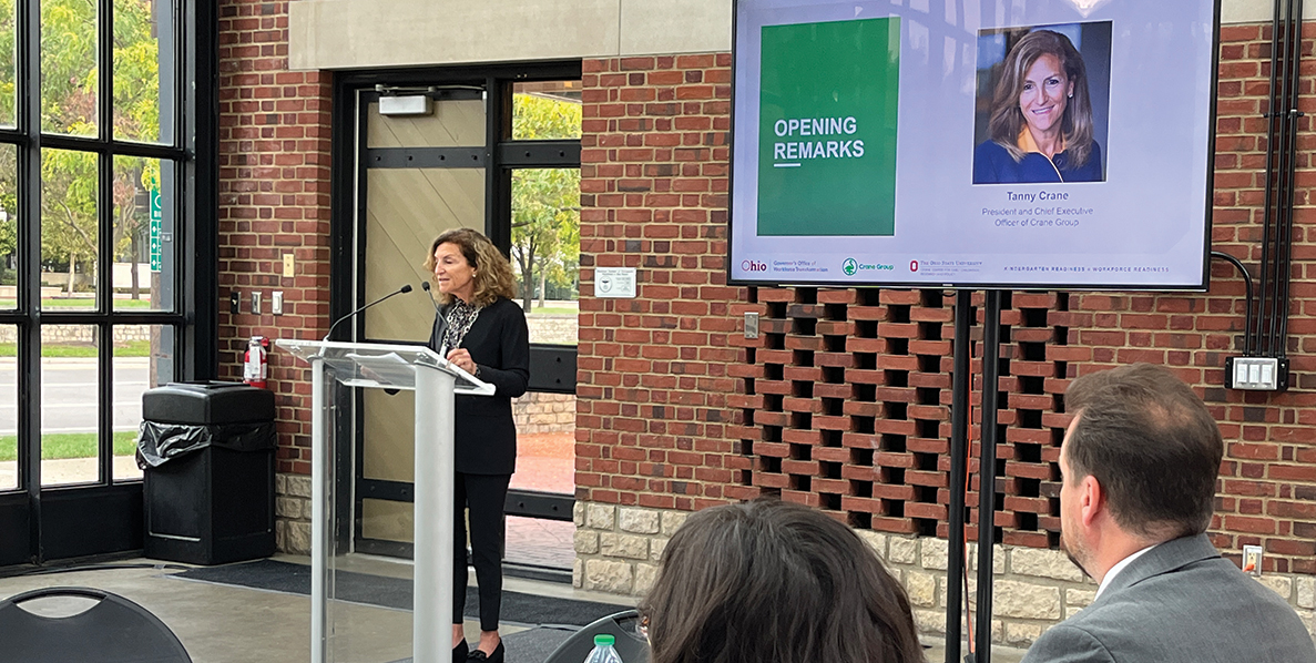 Tanny Crane, President and CEO of Crane Group, stands behind a podium and speaks into a pair of microphones attached to the podium. To the right is a video screen with a thumbnail photo of Crane next to a green box on which are white letters that read "OPENING REMARKS."