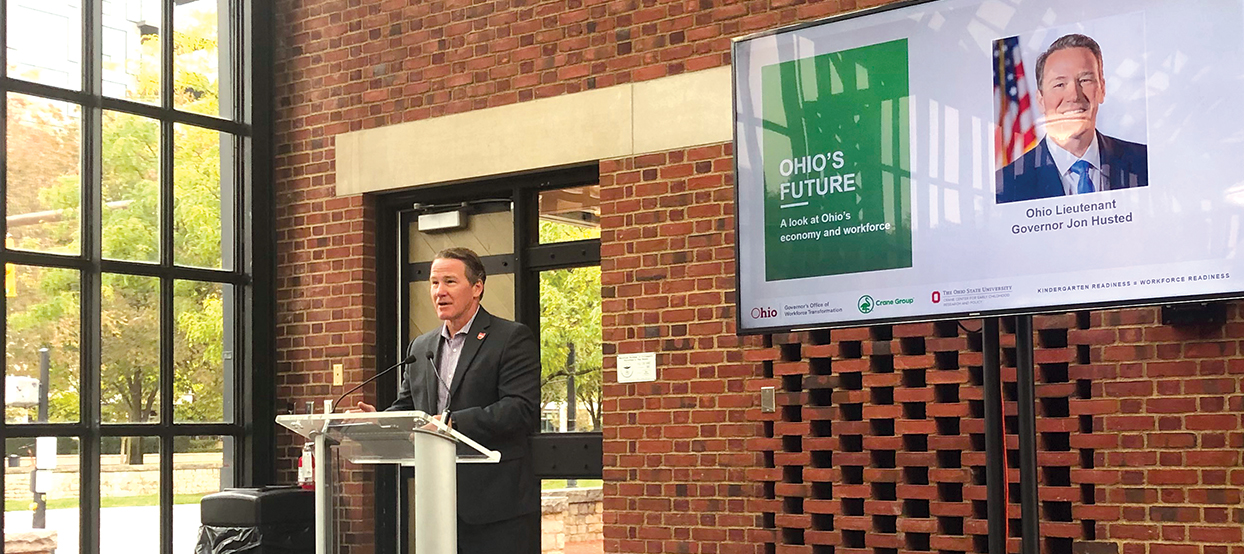 Ohio Lieutenant Governor Jon Husted stands behind a podium addressing the audience. To the right is a video screen showing a headshot photo of Husted next to a green box over which are white letters that read "OHIO'S FUTURE: A look at Ohio's economy and workforce."