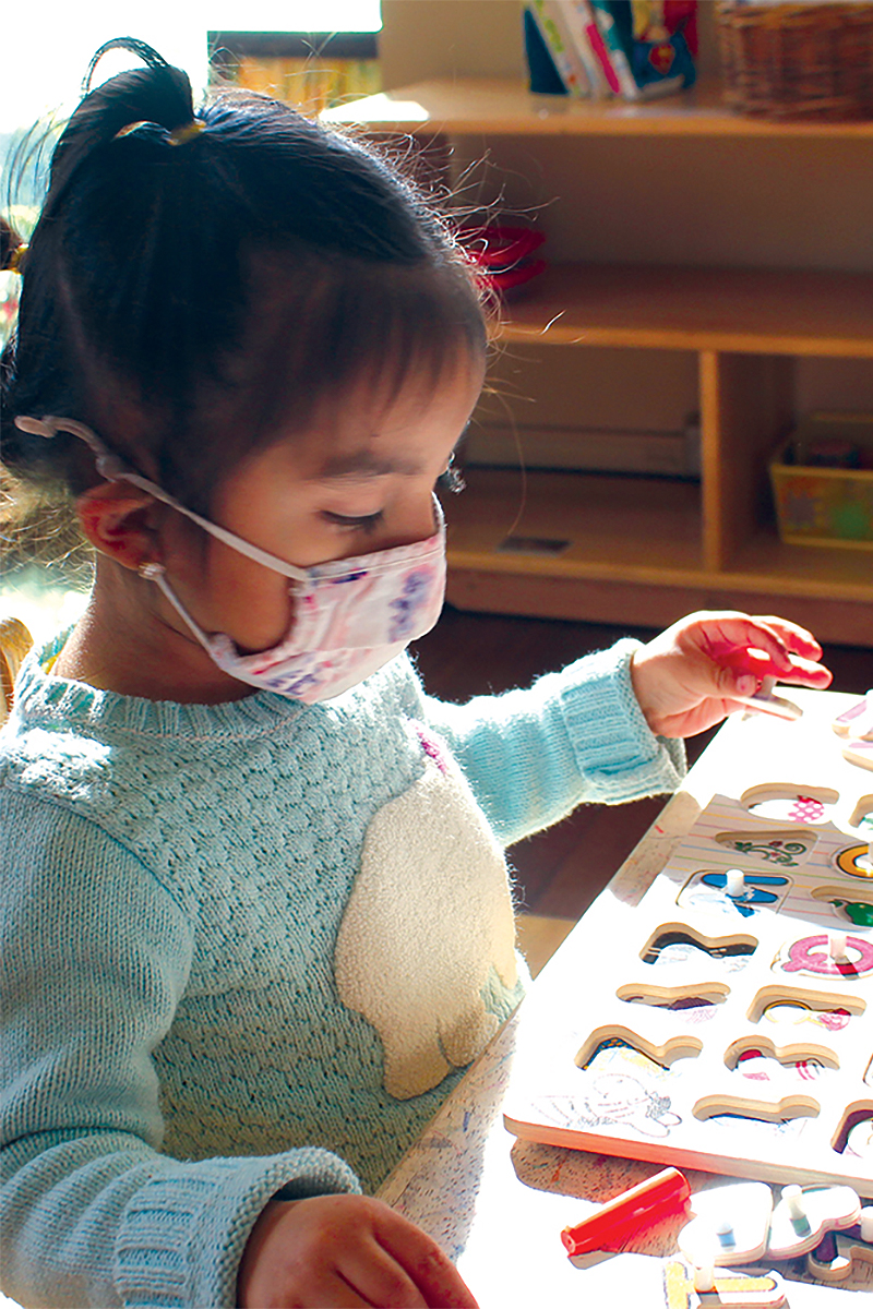 A small child wearing a tie-dye fabric face mask works on a wooden puzzle with cutouts of the letters of the alphabet.