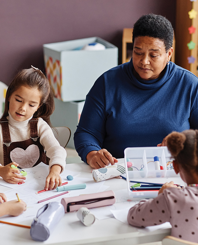 A preschool teacher sits at a table with students using colored pencils and markers to make drawings on sheets of white paper.