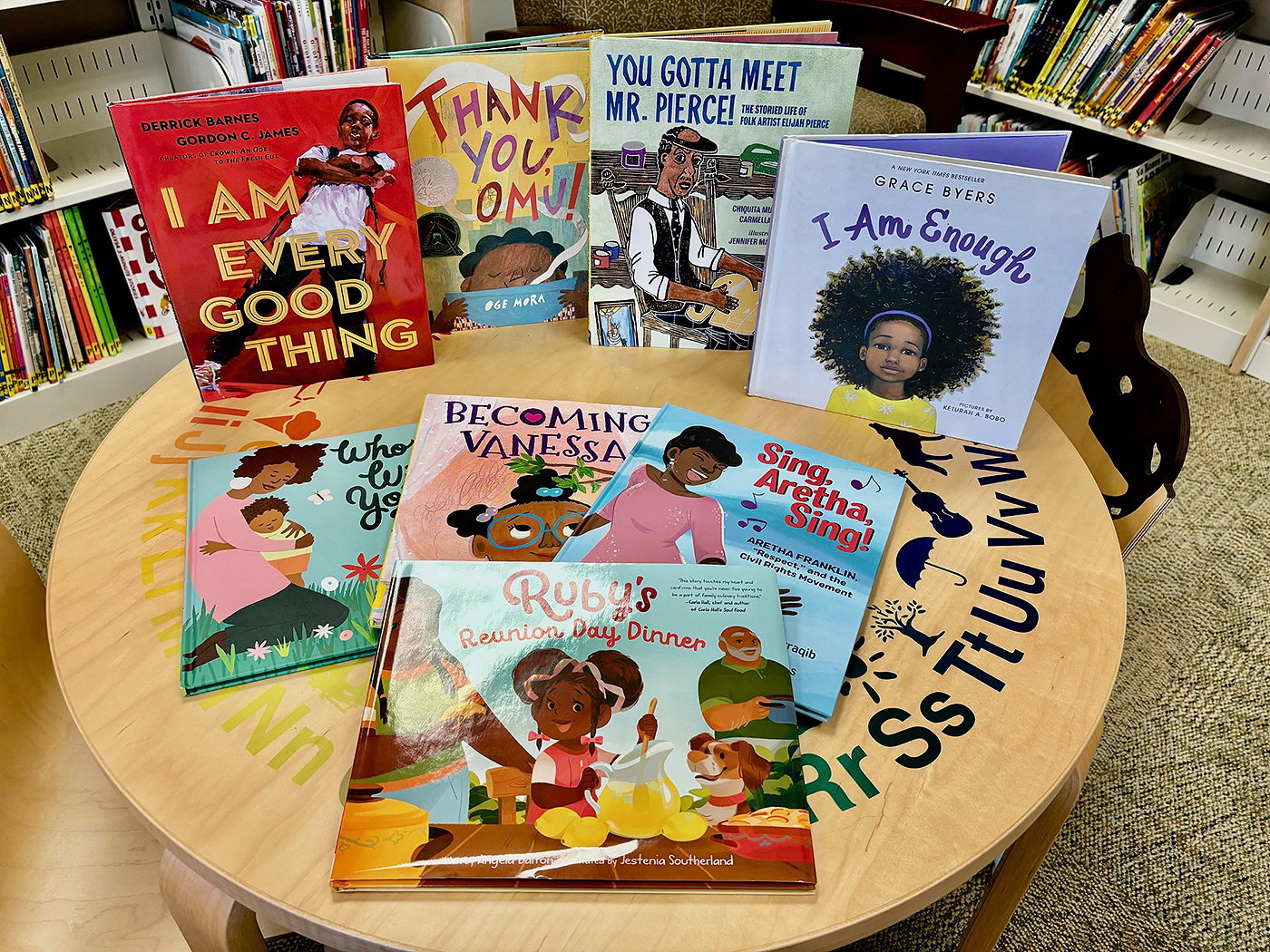 On top of a round table are eight children’s books chosen to commemorate Black History Month. Among the book titles are “Sing, Aretha, Sing,” “Ruby’s Reunion Day Dinner,” and “You Gotta Meet Mr. Pierce!”