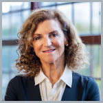 Headshot of Tanny Crane in front of windows. She smiles at the camera, has curly hair, and is wearing a white collared shirt and blue jacket.