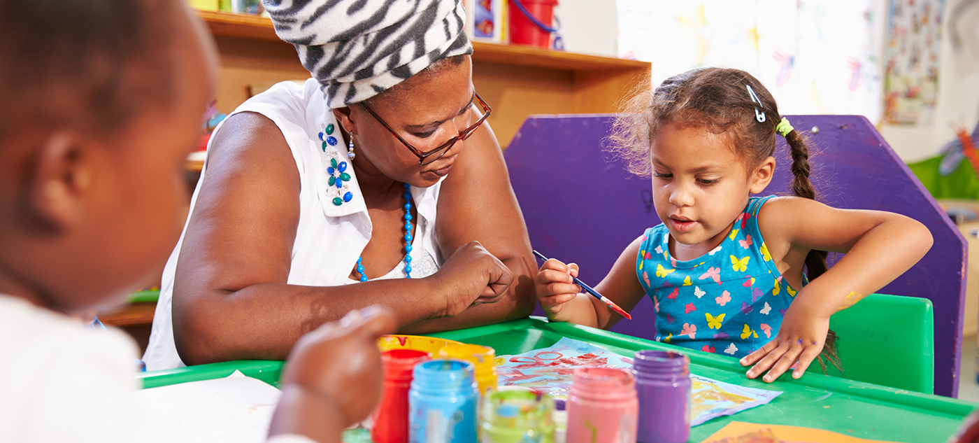 Close-up of a teacher sitting at a table with students. Teacher is wearing a zebra-print head covering and the girl she is talking to is wearing a blue tank top.