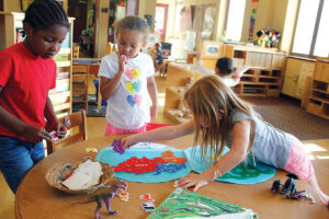 Three young children stand around a circular table. One child places a paper cutout in the shape of the continent of Australia on a round light blue circle representing the eastern hemisphere. The continents of Asia, Africa, and Europe are already in place on the light blue circle. Each of the continent cutouts displays the name of the continent.