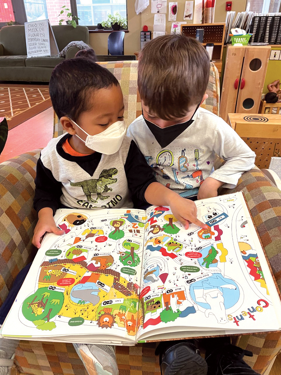 Two young children sit together in an upholstered chair looking at a large open book resting on their legs. The pages of the book display various images with words defining each image. One child points to an image on one of the open pages.