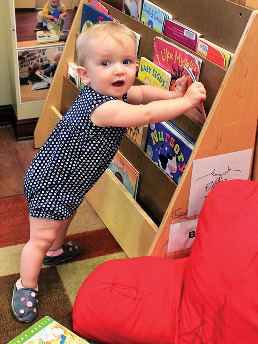 A toddler looks to the camera while grasping with both hands a children's book in a book rack.