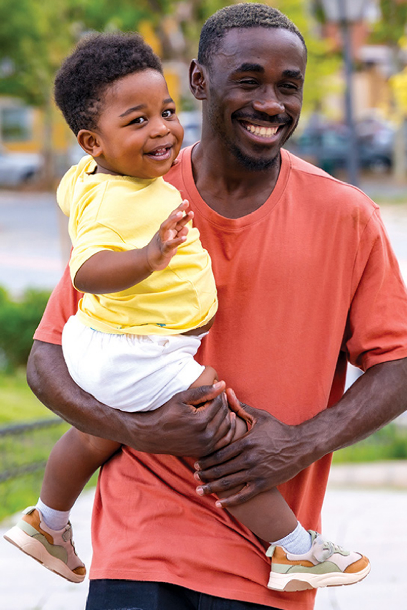 A man wearing an orange t-shirt holds a toddler boy in his arms as they walk into a park. The toddler is wearing a yellow t-shirt and white shorts, along with white socks and tennis shoes that are tan, olive and orange. Both the toddler and the man are looking at the camera and smiling.