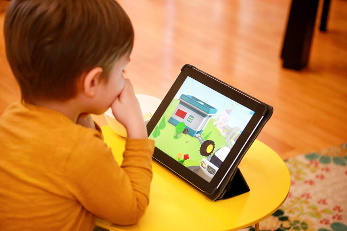 A small child is seated at a small table watching on a tablet computer an animated scene showing a building, a truck, grass, trees, and flowers.