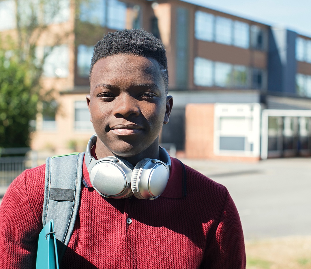 A teenage boy in a red shirt stands outside of his school building and smiles at the camera. The sunlight is bright and he has a folder under his arm, book bag on one shoulder, and headphones around his neck.