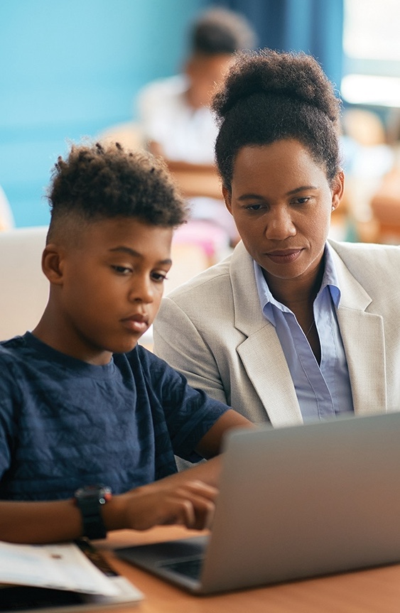 A female researcher in a white blazer sits next to a middle school boy in a blue shirt. Both are looking at his computer.