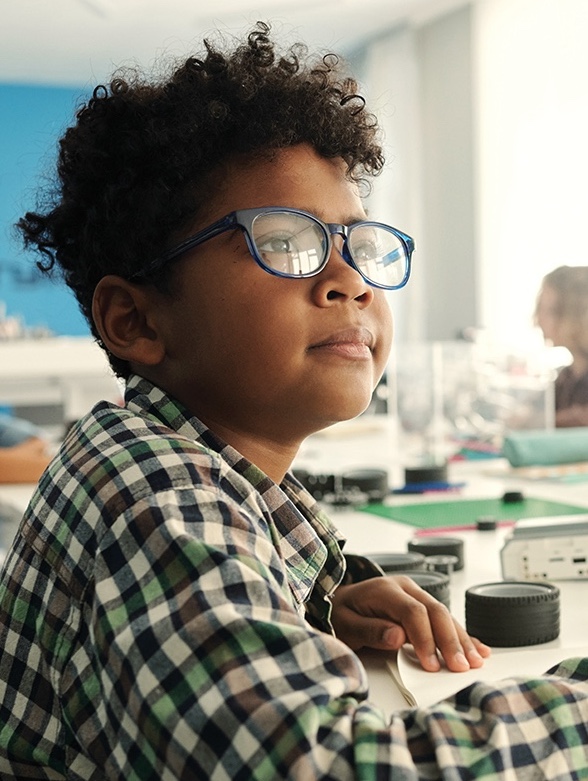 A middle school boy is seated at a large table in what appears to be a science classroom. The boy, who is wearing black-framed glasses, is looking to the right of the camera. The boy is wearing a black, brown, white and green plaid flannel long-sleeved shirt. On the table are small wheels and scientific equipment.