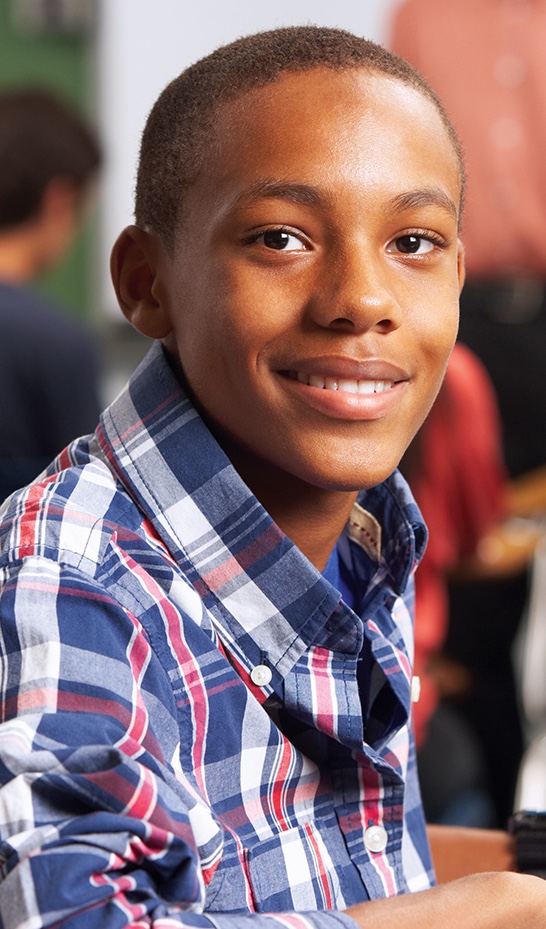 A middle school boy looks at the camera and smiles. He is wearing a red, white, and blue plaid button-down shirt over a blue t-shirt.