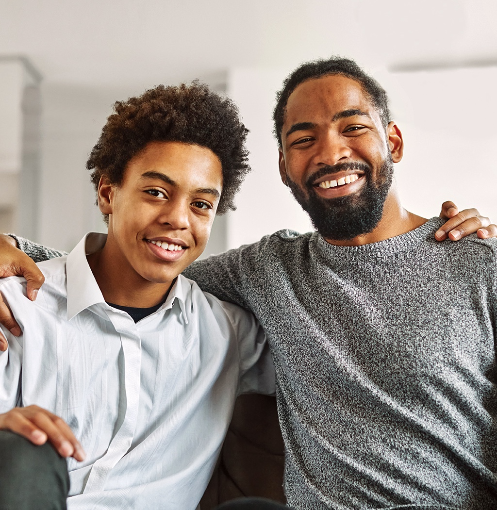 A father and his teenage son sit beside each other on a couch. They each have their inner arm wrapped around the other person's shoulders and are smiling at the camera.