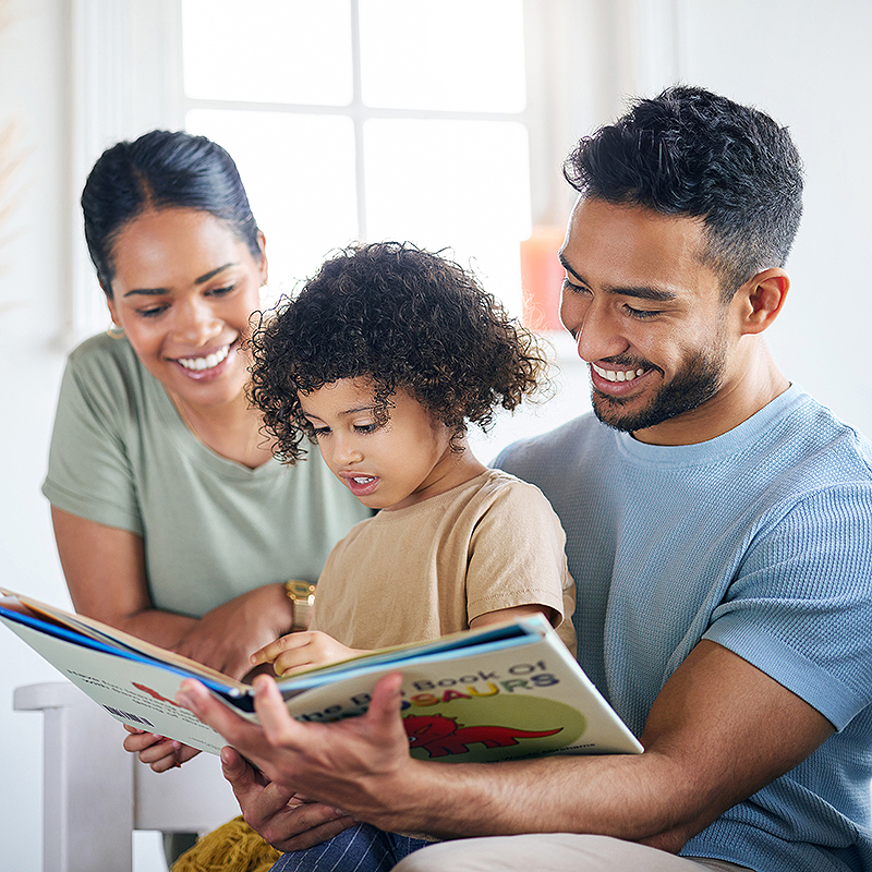 A man rests a young child on his knee, his arms wrapped around an open children's book titled "The Big Book Of Dinosaurs." The child points to something inside the open book, while a woman looks on over the man's right shoulder. The man and the woman are both smiling as the child appears to talking about something in the book.