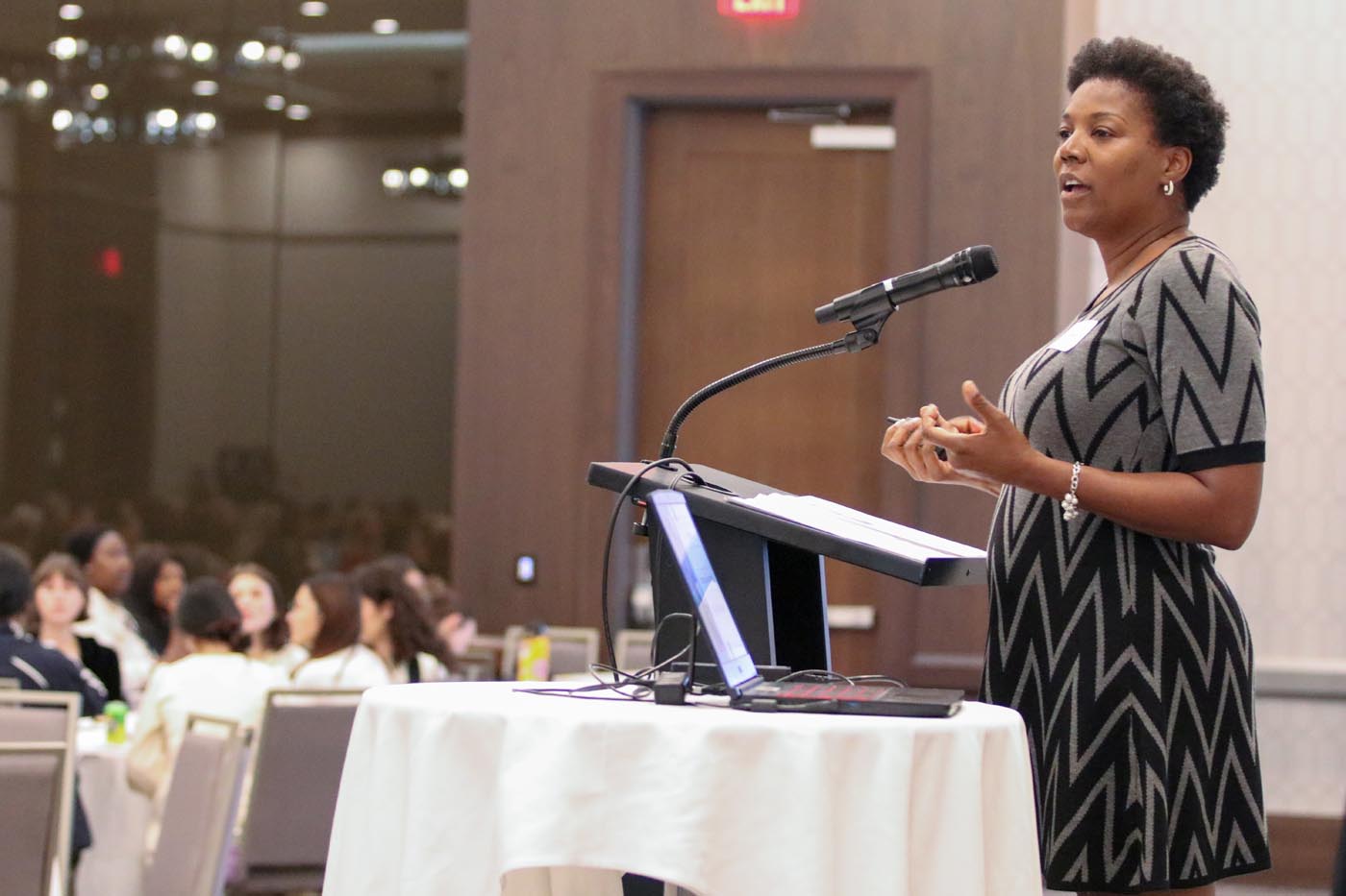 Tonya Coston speaks from the podium to the audience in a large hotel conference meeting room.