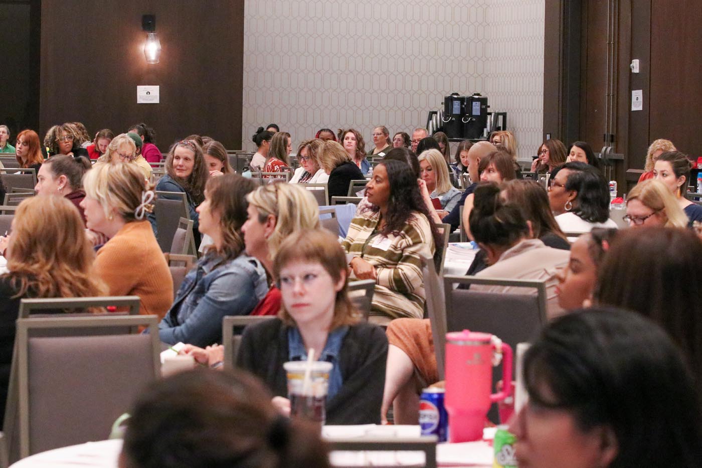 A crowd of people sit at round tables in a large, hotel conference room listening to a talk.