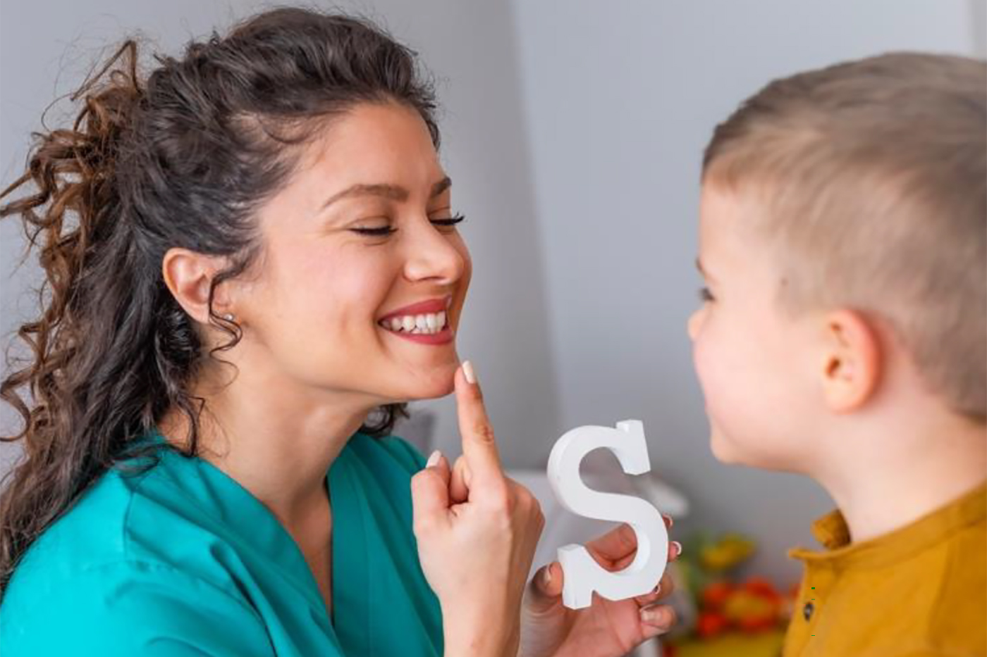 A speech pathologist points to her mouth while holding a painted wooden letter S. A child is looking at the mouth of the speech pathologist as the letter S is pronounced.