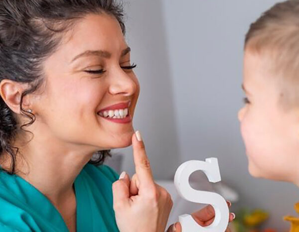 A speech pathologist points to her mouth while holding a painted wooden letter S. A child is looking at the mouth of the speech pathologist as the letter S is pronounced.